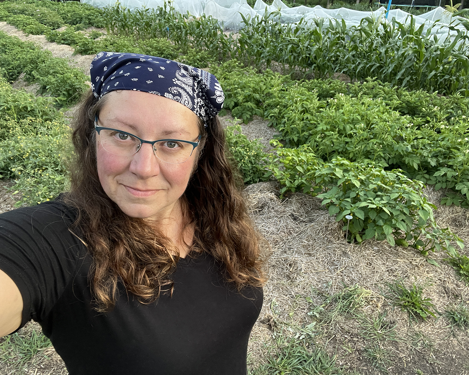 Kim Ross wearing a bandana and a black shirt smiling up at the camera from beside her no-till garden beds in summer time