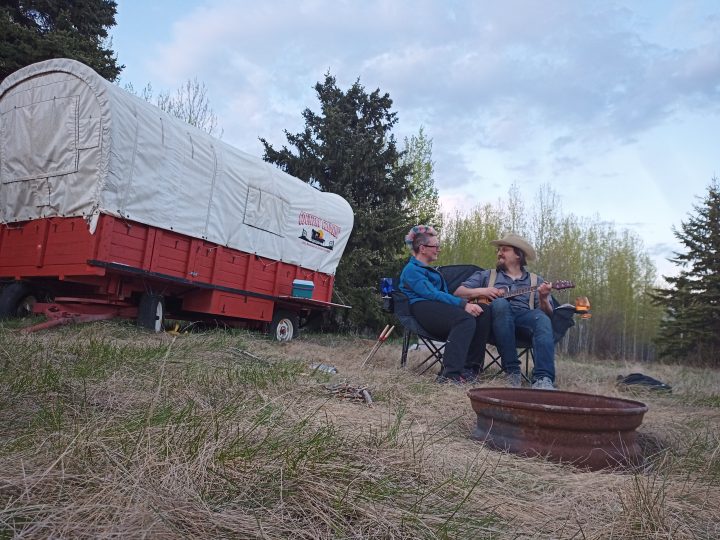 Anton Goulko and Dorothy Penner sitting in chairs by an outdoor fire in a field on their farm, beside their farm stay caravan. 