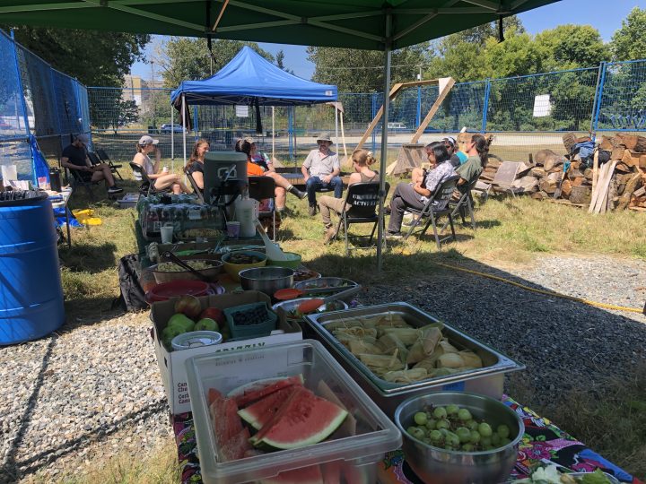 Food in holders of many shapes and sizes is packed along two folding tables, under foldable shade tents. Watermelon, grapes and tamales in the foreground, stretching back to flats of juice and a large cooler of horchata at the far end. Beyond the table 10 or so folks sit on folding chairs, in circle, in the sun. There is a blue tent offering shade, and a blue fence surrounding the work area.