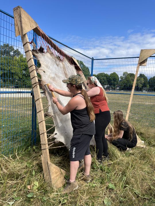 Three folks are scraping a raw hide, stretched tightly with jute roping, on an 8 x 8 woodern frame that has warped with time and so is bent and uneven. Two are standing and using long scrapers and one is kneeling at work. The park in the distance is wide open, grass and a sand softball pitch are visible, with maples and cottonwoods green in the distance, and a big open blue sky above with some low lying cloud.