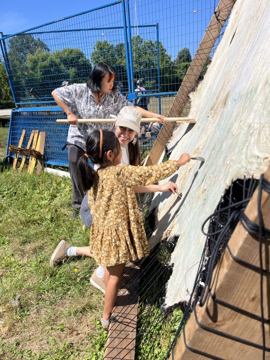 A young one works with their friend to feel the scraping process and contribute to softening the hide. The young one stands in a floral dress, and the friend kneels to demonstrate how to hold the tool. Another person works with a wooden dowel in the background.