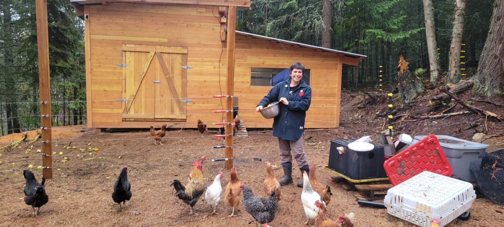 A young woman with short, brown hair standing outside a new barn holding a shallow feeding pail surrounded by chickens.